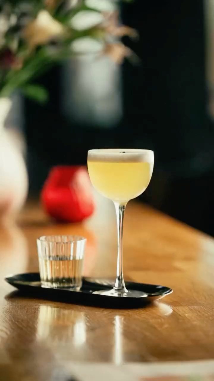 A frothy coupe cocktail on the bar with flowers behind