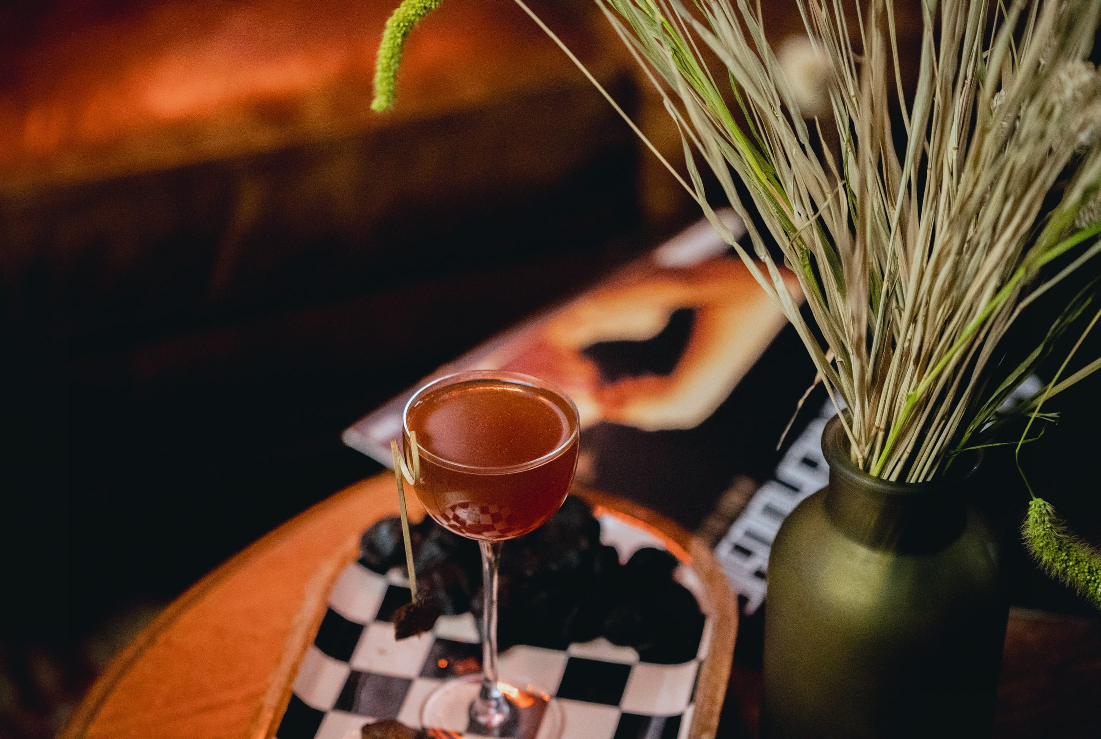 An amber cocktail in a nick-and-nora glass on a checkered tray beside dried grasses