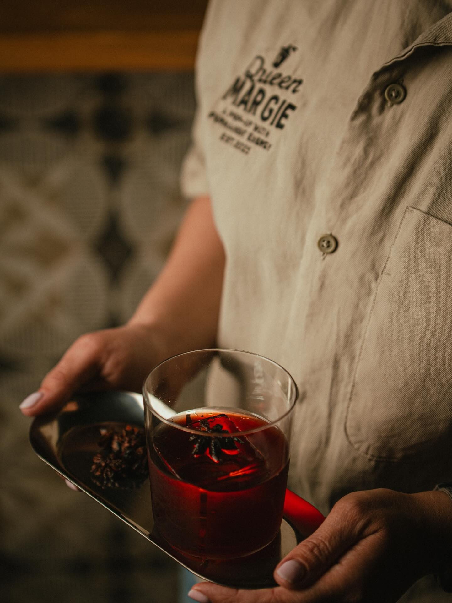A red cocktail with a star anise garnish, held on a steel tray